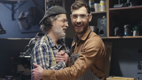 Portrait-shot-of-two-cheerful-father-and-son-in-goggles-hugging-and-shaking-hands-in-workshop
