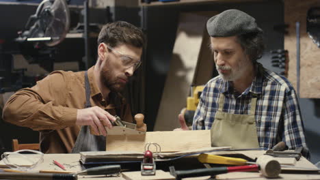 Old-father-carpenter-teaching-his-young-son-to-process-and-work-with-timber-in-workshop