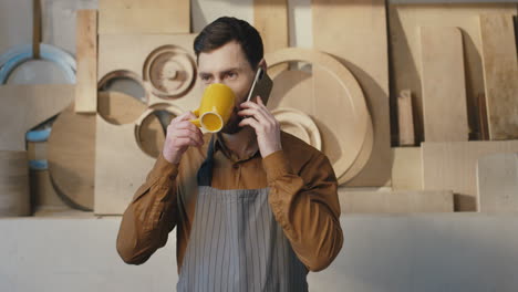Portrait-of-caucasian-bearded-man-in-apron-talking-on-the-phone-and-drinking-coffee-in-carpentry-workshop