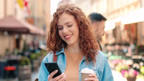 Redheaded-woman-holding-coffee-to-go-and-using-smartphone-in-the-street