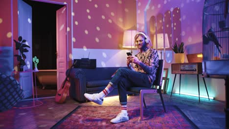 Young-stylish-caucasian-man-with-long-hair-in-headphones-sitting-on-a-chair-and-using-smartphone-in-a-retro-party-at-home