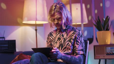 Young-stylish-caucasian-man-with-long-hair-in-headphones-sitting-on-a-chair-and-using-tablet-in-a-retro-party-at-home