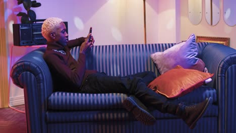 Young-stylish-african-american-sitting-on-sofa-and-chatting-online-with-friends-using-smartphone-while-waiting-for-the-party-to-start