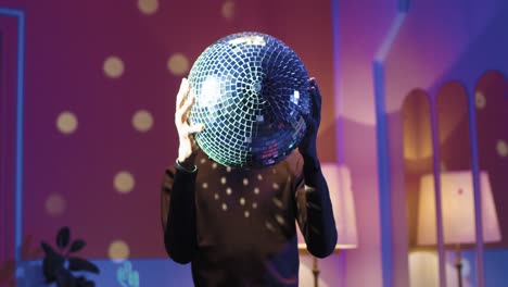 Young-stylish-man-holding-disco-ball-in-his-hands-standing-statically-in-a-retro-party-at-home