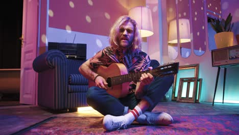 Young-stylish-caucasian-man-with-long-hair-sitting-on-the-floor-and-playing-the-guitar-in-a-retro-party-at-home