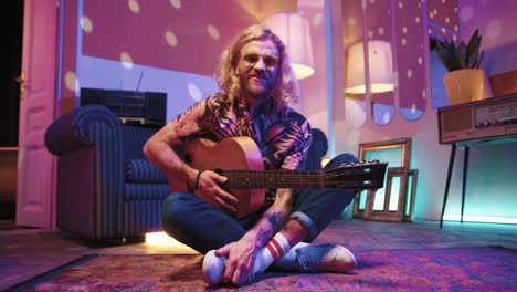 Young-stylish-caucasian-man-with-long-hair-sitting-on-the-floor-and-playing-the-guitar-in-a-retro-party-at-home