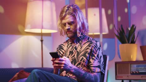 Young-stylish-caucasian-man-with-long-hair-in-glasses-sitting-on-a-chair-and-using-smartphone-in-a-retro-party-at-home