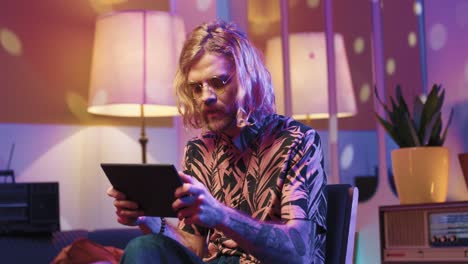 Young-stylish-caucasian-man-with-long-hair-in-glasses-sitting-on-a-chair-and-using-a-tablet-in-a-retro-party-at-home