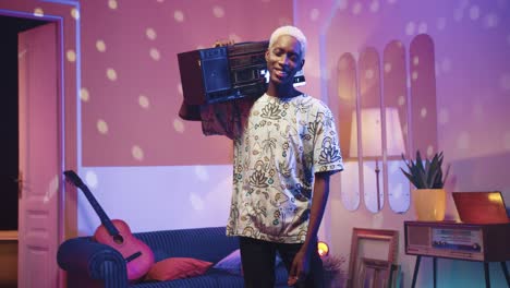 Happy-cheerful-young-African-American-guy-standing-in-living-room-in-a-retro-party-at-home-holding-retro-boom-box-on-a-shoulder-and-looking-at-camera