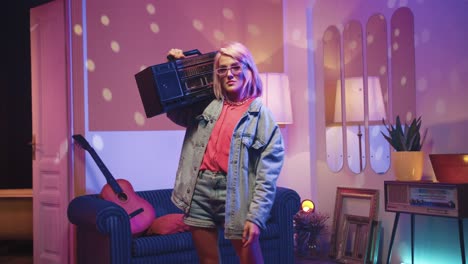 Happy-cheerful-young-caucasian-woman-in-glasses-standing-in-living-room-in-a-retro-party-at-home-holding-retro-boom-box-on-a-shoulder-and-looking-at-camera