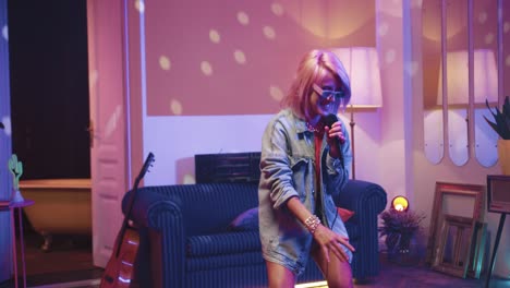 Joyful-Caucasian-young-woman-with-blonde-hair-and-glasses-singing-in-microphone-and-dancing-in-a-retro-party-at-home