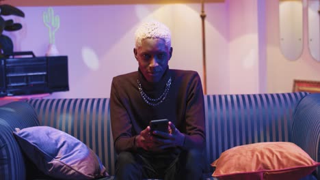 Portrait-of-happy-African-American-young-smiling-man-with-blonde-hair-typing-on-smartphone-and-looking-at-camera-while-sitting-on-a-sofa-in-a-retro-room-at-home