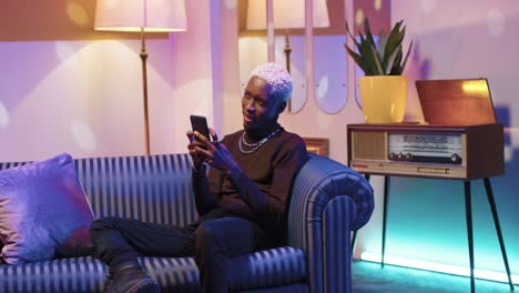 Afroamerikanischer-Mann-Mit-Blonden-Haaren-Benutzt-Smartphone,-Während-Er-Zu-Hause-Auf-Dem-Sofa-In-Einem-Retro-zimmer-Sitzt