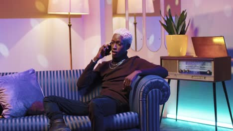 African-american-man-with-blonde-hair-talking-on-the-smartphone-while-sitting-on-sofa-in-a-retro-room-at-home
