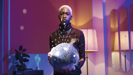 Young-stylish-man-holding-disco-ball-in-his-hands-and-looking-at-camera-in-a-retro-party-at-home