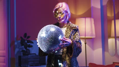 Young-stylish-woman-in-glasses-holding-disco-ball-in-her-hands-and-looking-at-camera-in-a-retro-party-at-home