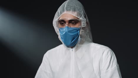 Caucasian-male-doctor-in-Personal-Protective-Equipment-and-medical-mask,-putting-on-glasses-and-looking-at-side-on-black-wall-background