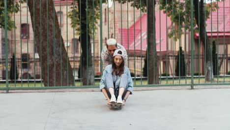 Full-length-view-of-a-teenage-girl-sitting-on-a-skateboard-while-her-african-american-boyfriend-pushing-her-in-the-street