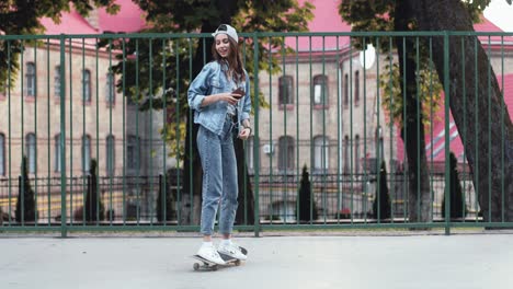 Full-length-view-of-a-teenage-girl-listening-music-with-smartphone-while-standing-on-her-skateboard-and-dancing-in-the-street