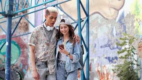 Smiling-man-and-his-caucasian-girlfriend-wearing-headphones-and-watching-something-on-the-smartphone-while-walking-down-the-street