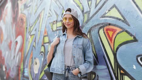 Portrait-shot-of-young-caucasian-stylish-hipster-girl-standing-at-graffity-wall-and-smiling-at-the-camera