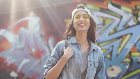 Portrait-shot-of-young-caucasian-stylish-hipster-girl-standing-at-graffity-wall-and-smiling-at-the-camera