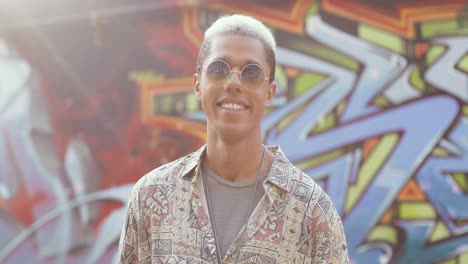 Portrait-shot-of-young-caucasian-stylish-hipster-man-standing-at-the-graffity-wall-and-smiling-at-the-camera