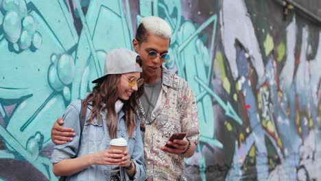 Hipster-paar-In-Stylischer-Kleidung,-Das-Etwas-Auf-Dem-Smartphone-In-Der-Nähe-Einer-Grafitti-wand-Auf-Der-Straße-Beobachtet