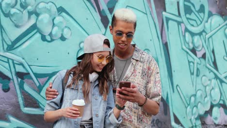 Hipster-couple-in-stylish-clothes-watching-something-on-smartphone-near-a-grafitti-wall-in-the-street
