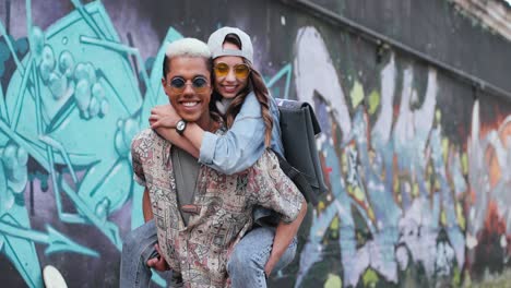 Young-boy-giving-her-girlfriend-a-piggyback-and-looking-at-the-camera-near-a-graffity-wall-in-the-street