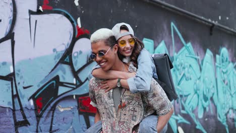 Young-boy-giving-her-girlfriend-a-piggyback-and-walking-near-a-graffity-wall-in-the-street