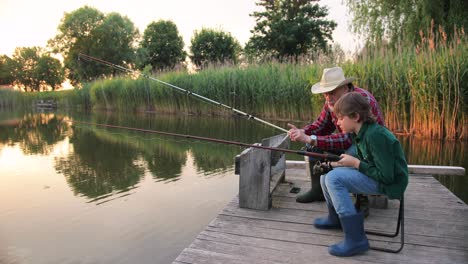 Side-view-of-a-teen-boy-sitting-with-his-grandfather-on-the-lake-pier,-talking-and-holding-a-fish-on-the-fishing-rods-while-fishing-together