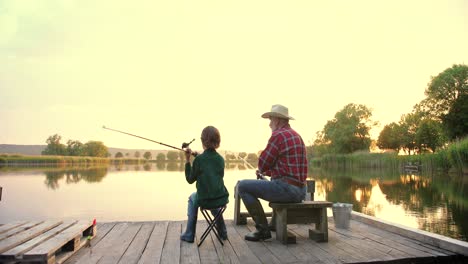 Rear-view-of-a-teen-boy-sitting-with-his-grandfather-on-the-lake-pier,-talking-and-fishing-together