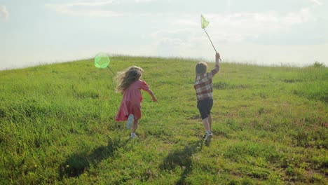 Rear-view-of-two-happy-children-holding-a-net-for-catching-insects-running-through-the-green-field
