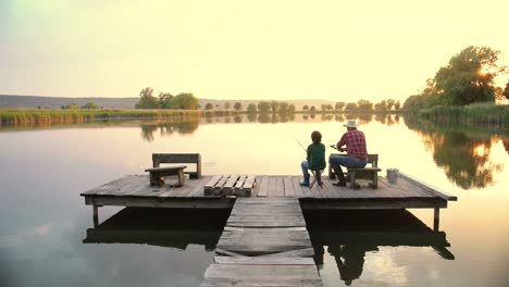 Rear-view-of-a-teen-boy-sitting-with-his-grandfather-on-the-lake-pier,-talking-and-fishing-together