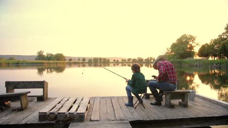 Camera-zooming-from-the-back-of-a-teen-boy-sitting-with-his-grandfather-on-the-lake-pier,-talking-and-fishing-together