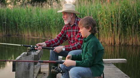 Side-view-of-a-teen-boy-sitting-with-his-grandfather-on-the-lake-pier-fishing-together