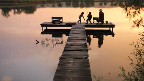 Distant-rear-view-of-a-teen-boy-and-girl-sitting-with-their-grandfather-on-the-lake-pier-and-fishing-together