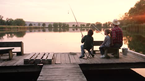 Camera-zooming-from-the-back-of-a-teen-boy-and-girl-sitting-with-their-grandfather-on-the-lake-pier-and-fishing-together