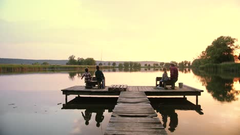 Distant-rear-view-teen-boys-and-girl-sitting-with-their-grandfather-on-the-lake-pier-and-fishing-together