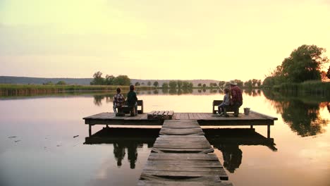 Distant-rear-view-teen-boys-and-girl-sitting-with-their-grandfather-on-the-lake-pier-and-fishing-together