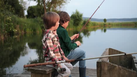 Side-view-of-two-teen-boys-sitting-on-the-lake-pier,-talking-and-fishing-together