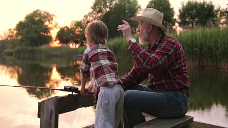Side-view-of-a-grandfather-and-his-grandson-fishing-sitting-on-the-lake-pier-on-a-summer-day-at-sunset
