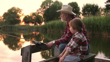 Close-up-view-of-a-grandfather-and-his-grandson-fishing-and-talking-sitting-on-the-lake-pier-on-a-summer-day-at-sunset