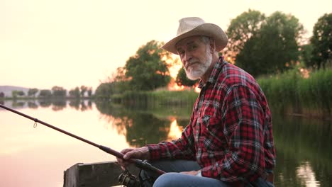 Portrait-of-a-senior-fisherman-wearing-hat-and-sitting-on-the-lake-pier-while-holding-a-fishing-rod-at-sunset