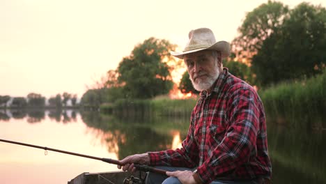 Portrait-of-a-senior-fisherman-putting-on-hat-and-sitting-on-the-lake-pier-while-holding-a-fishing-rod-at-sunset