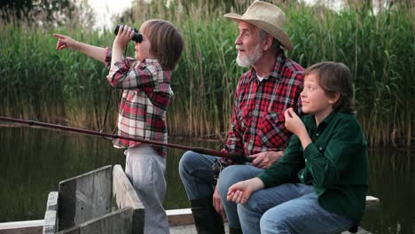 Cute-little-boy-using-binoculars-and-showing-something-to-his-grandfather-and-his-brother-while-they-are-fishing-sitting-on-the-lake-pier