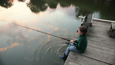 High-angle-view-of-a-little-boy-fishing-while-he-is-sitting-on-the-lake-pier-at-sunset
