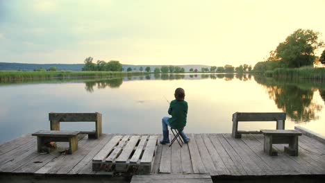 Rear-view-of-a-teen-boy-sitting-on-the-lake-pier-and-fishing-at-sunset