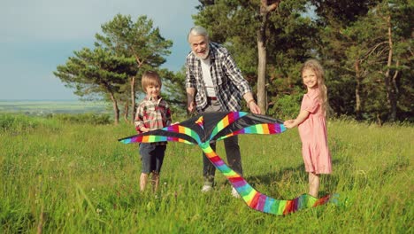 Caucasian-senior-man-with-his-grandchildren-holding-a-kite-and-looking-at-the-camera-in-the-park-on-a-sunny-day